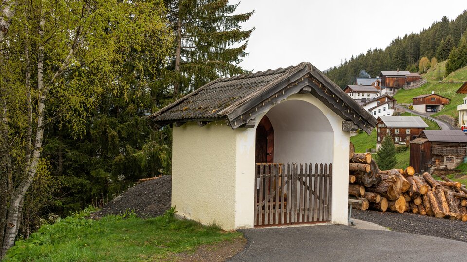 A small, charming cottage with an angled roof stands by a path. In the background, there are wood piles and some houses in a wooded mountain landscape. | © Bernhard Gruber – Photography