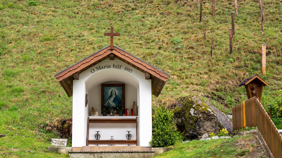 A small chapel with a cross and a picture of the Virgin Mary. It stands in the middle of a green meadow.