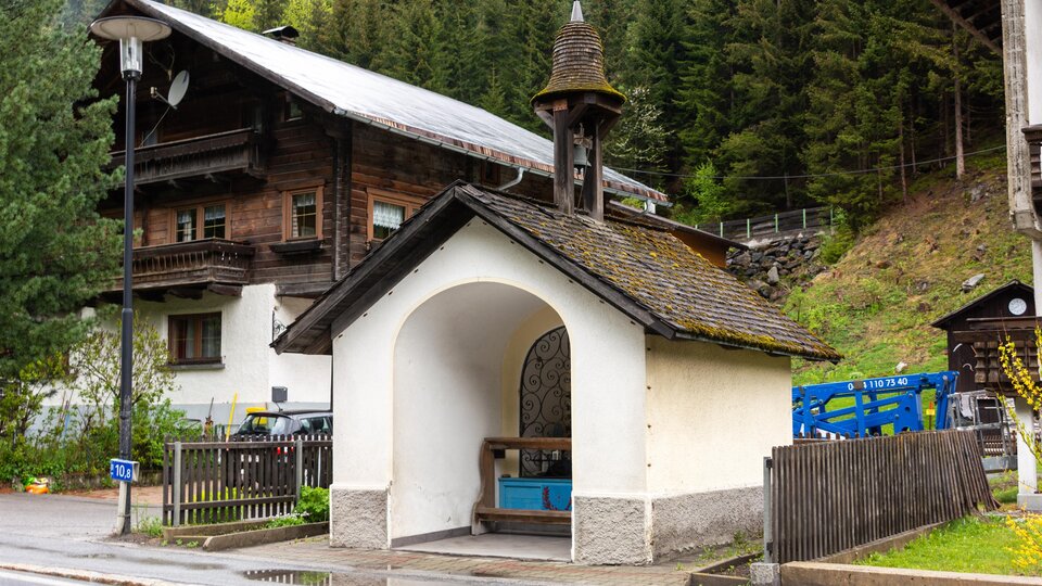 A small white chapel with a bell tower stands by the road. In the background, typical alpine wooden houses and trees can be seen. | © Bernhard Gruber – Photography