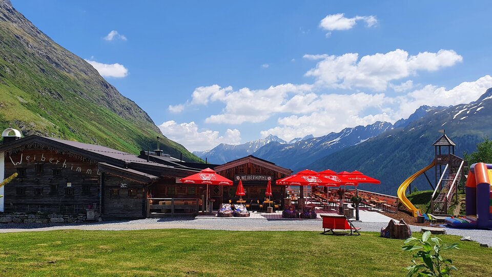 A cozy inn in the mountains with red sun umbrellas. In the background, green meadows and majestic mountains can be seen.