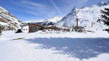 A picturesque mountain landscape covered in snow with a small building and people outdoors. In the background, impressive mountains are visible.