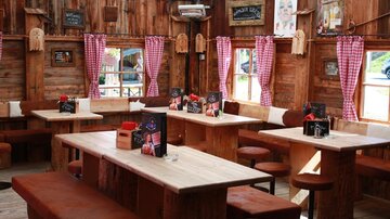 A cozy restaurant with wooden paneling and rustic tables. The windows are decorated with checked curtains and there are signs on the walls.