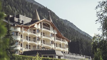 An elegant hotel with towers, surrounded by green trees and mountains. The architecture combines modern and alpine design elements. | © Hannes Niederkofler