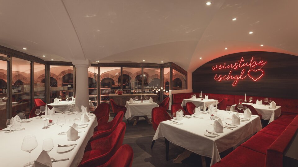 A stylish, warmly lit wine cellar with elegantly set tables, red velvet armchairs, and a glowing neon sign "Weinstube Ischgl" creates a cozy-exclusive atmosphere. | © Hannes Niederkofler