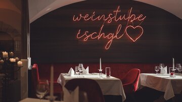 A stylish restaurant area with dressed tables, red velvet seats, and a warmly glowing neon sign "Weinstube Ischgl" conveys an elegant, cozy atmosphere. | © Hannes Niederkofler