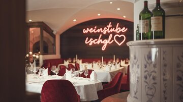 An elegant restaurant area with white-covered tables, red velvet chairs, and the warmly glowing neon sign "Weinstube Ischgl" creates a stylish, cozy atmosphere. | © Hannes Niederkofler