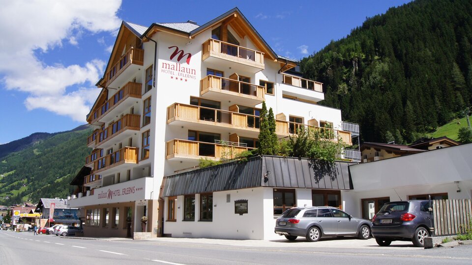 A modern hotel building with several floors and balconies. Surrounded by green mountains and clear sky.
