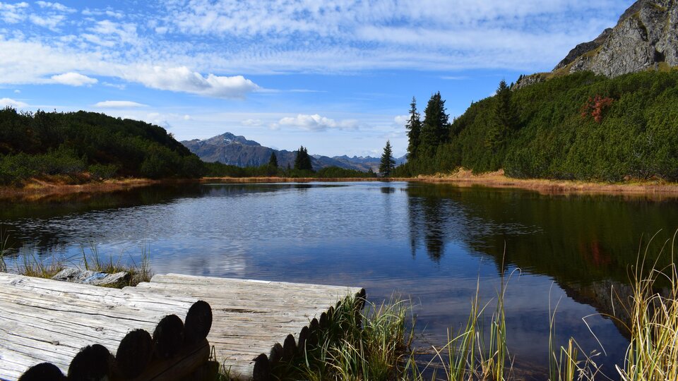A tranquil lake surrounded by mountains and forest. The sky is clear with a few clouds.
