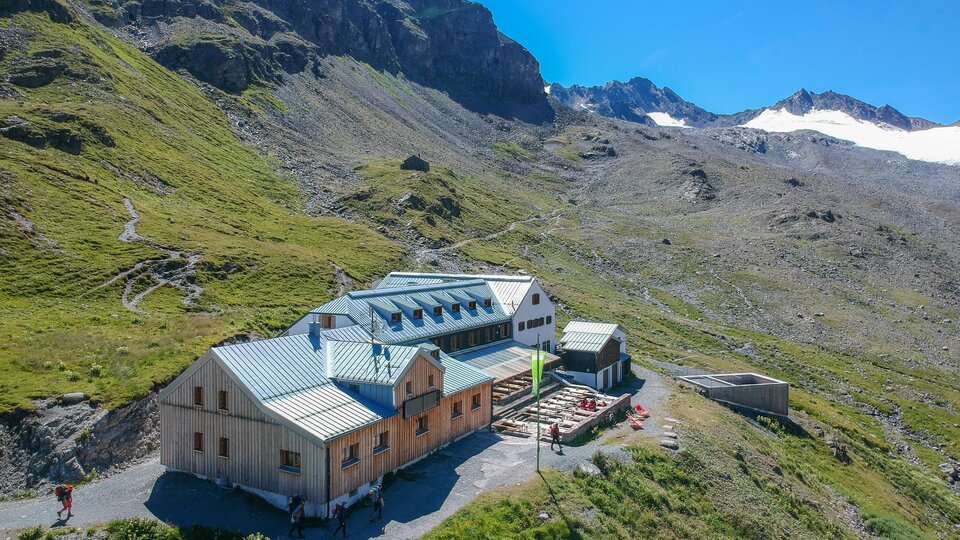 A mountain cabin surrounded by green meadows and mountains. The sky is clear and blue, perfect for an outdoor vacation.
