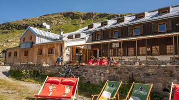 A cozy mountain house with a wooden facade and a terrace. Colorful loungers are in the foreground, and several people are enjoying the view.