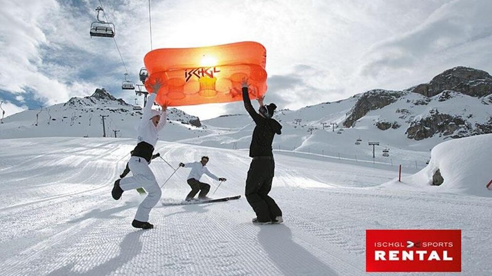 A group of skiers is standing in the snow, holding an orange umbrella above them. In the background, there are snow-covered mountains and a chairlift.