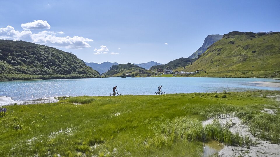 Two cyclists ride along a sparkling lake. In the background, green hills and a blue sky are visible.