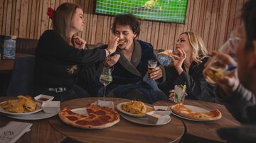 A group of friends is sitting in a restaurant enjoying food and drinks. In the background, a football game is playing on television.