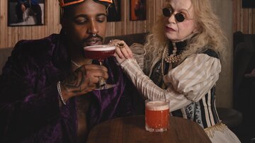 An elegant bar with two people. A woman hands a drink to a man while they are wearing stylish clothing.