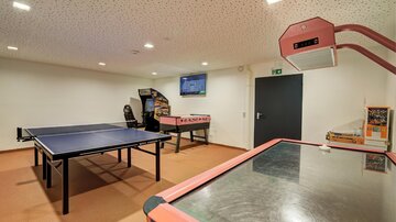 Ein Raum mit einem Tischtennistisch und einem Airhockey-Tisch. Im Hintergrund steht ein Fernseher und es gibt weitere Spielgeräte.