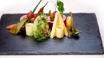 Ein kunstvoll angerichteter Teller mit verschiedenen Gemüsesorten, Käse und kleinen Beilagen. Die Präsentation erfolgt auf einer schwarzen Platte.