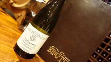 Eine Flasche Wein mit einem weißen Etikett steht auf einem Holztisch. Im Hintergrund ist ein Menü oder eine Weinkarte zu sehen.