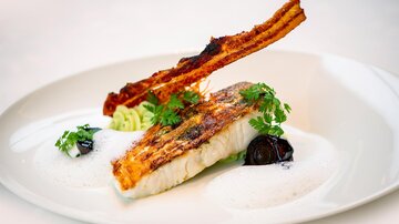 Ein fein angerichteter Teller mit gebratenem Fisch, garniert mit knusprigem Speck und frischen Kräutern. Dazu gibt es eine schaumige Sauce und schwarze Oliven.