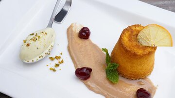 Ein leichtes Dessert bestehend aus einem bunten Küchlein, einer Kugel Eis und einer Creme. Es ist mit Kirschen und einem Zitronenschnitz garniert.