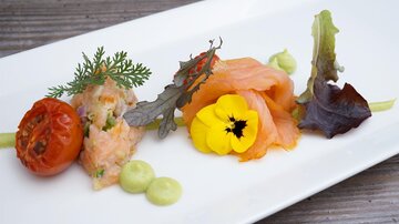 Ein ansprechend angerichtetes Gericht mit Lachs, einem gebratenen Tomaten und frischen Kräutern. Dekorative Blüten und grüne Saucen sorgen für einen Farbenfrohen Akzent.