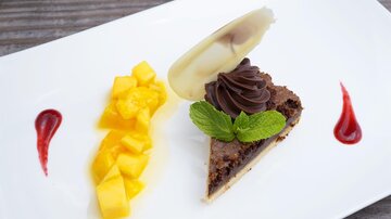 Ein Stück Schokoladenkuchen mit einer dekorativen Sahnehaube und einem Blatt Minze. Neben dem Kuchen befinden sich frische Mangowürfel und Fruchtsoße.