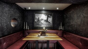 Ein stilvolles Restaurant mit einem gemütlichen Sitzbereich in dunklen Tönen. An der Wand hängt ein Schwarz-Weiß-Bild, und der Tisch ist mit einer kleinen Dekoration geschmückt.