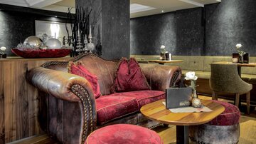 Eine stilvolle Lounge mit einem roten Samtsofa und einem runden Tisch. Die Wände sind dunkel und dezent dekoriert, was eine gemütliche Atmosphäre schafft.