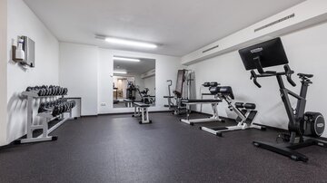 Ein schlichtes Fitnessstudio mit verschiedenen Trainingsgeräten. Es gibt Gewichte, eine Bank und ein Fahrradergometer in einem modernen Raum.