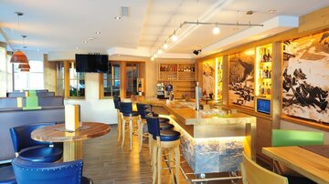 A modern interior with wooden paneling and stylish furniture. The bar is well-equipped and the room has large windows with plenty of natural light.