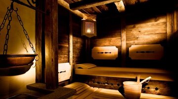 A cozy sauna area with warm wood and soft lighting. In the foreground, a sauna ladle and a bucket are visible.