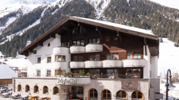 Ein charmantes Hotel im alpinen Stil, umgeben von schneebedeckten Bergen. Der Eingangsbereich zeigt eine schöne Fassade mit großen Fenstern.