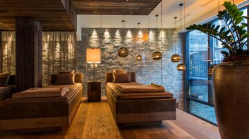 Ein modernes Spa mit gemütlichen Liegen und warmem Licht. Die Wände sind aus Stein und große Fenster bieten einen Blick nach draußen.