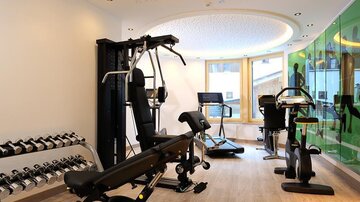 Ein modernes Fitnessstudio mit verschiedenen Trainingsgeräten, darunter Hanteln, eine Kraftstation und ein Ergometer. Der Raum ist hell und einladend mit großen Fenstern.