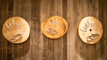 Drei runde Holzplatten hängen an einer Wand. Auf jeder Platte steht ein Wort in geschwungener Schrift: "Liebe", "Lust" und "Hunger".