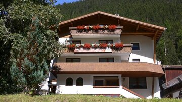 Ein charmantes Gebäude mit Balkonen, die mit blühenden Blumen dekoriert sind. Umgeben von Bäumen und einer malerischen Berglandschaft.