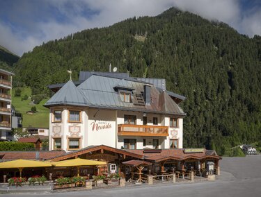 A charming hotel with a large balcony and an inviting terrace. In the background, green mountains and a cloudy sky can be seen.