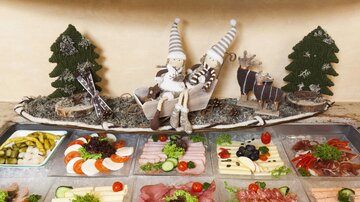 A lovely buffet with various sausage and cheese platters, garnished with fresh vegetables and decorations. In the background, festive figures and trees can be seen.