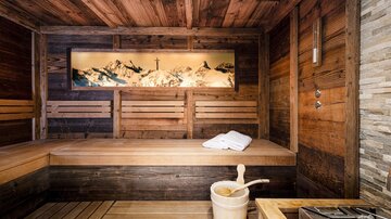 A cozy sauna with wooden cladding and a peaceful view of the mountains. A towel and a bucket are ready on the bench.