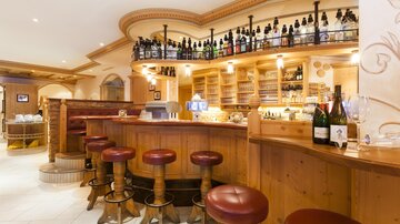 A cozy bar cellar with a wooden counter and tall bar stools. In the background, many bottles and glasses are on shelves.