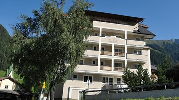 Ein modernes Gebäude mit mehreren Etagen und Balkonen. Umgeben von Bäumen und einer Berglandschaft im Hintergrund.