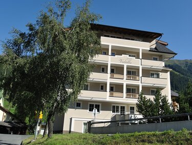Ein modernes Gebäude mit mehreren Etagen und Balkonen. Umgeben von Bäumen und einer Berglandschaft im Hintergrund.