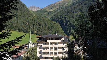 Eine malerische Berglandschaft mit grünen Wäldern und beeindruckenden Gipfeln. Im Vordergrund steht ein modernes Gebäude neben einer kleinen Kirche.