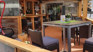 Ein gemütlicher Außenbereich eines Restaurants mit Holzdekor und modernen Möbeln. Bunte Blumen und eine Sonnenschutzmarkise sorgen für eine einladende Atmosphäre.