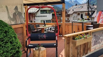 Eine gemütliche Terasse mit einem roten Sitzschaukelstuhl und schöner Holzverkleidung. Im Hintergrund sind die Berge und ein klarer blauer Himmel zu sehen.