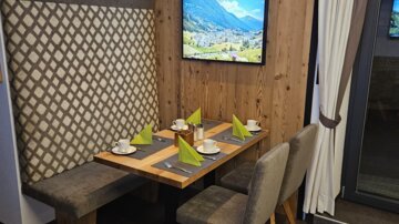 Ein gemütlicher Essbereich mit Holzdetails und einer modernen Sitzgelegenheit. Auf dem Tisch sind grüne Servietten und Tassen angeordnet, während im Hintergrund ein Fernseher mit Landschaftsbild zu sehen ist.