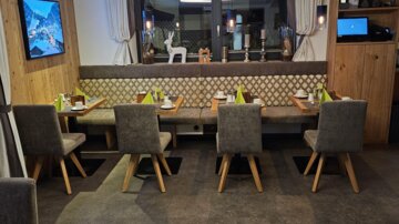 Ein modernes Restaurant mit Holzmöbeln und gemütlicher Atmosphäre. Es gibt mehrere Tische mit Stühlen und eine große Fensterfront.