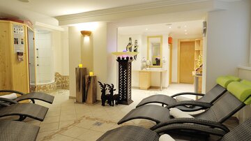 Ein entspannender Wellnessbereich mit Liegen, sanfter Beleuchtung und einem modernen Design. Im Hintergrund sind eine Dusche und eine Empfangstheke sichtbar.