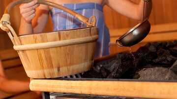 Eine Person gießt Wasser aus einer Kelle auf heiße Steine in einer Sauna. Daneben steht ein Holzkorb.