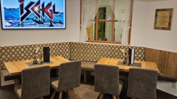Ein gemütlicher Restaurantbereich mit Holztischen und gepolsterten Stühlen. Im Hintergrund ist ein Fernseher mit dem Schriftzug "Ischgl" sichtbar.