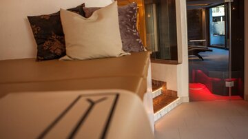 Ein stilvolles Wohnzimmer mit einer bequemen Couch und dekorativen Kissen. Im Hintergrund ist ein Raum mit sanfter Beleuchtung zu sehen.